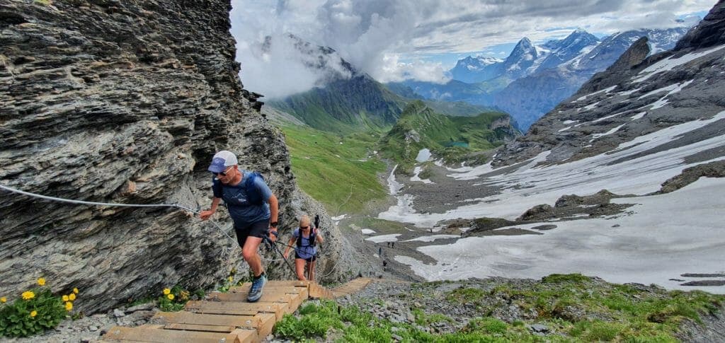 Male and female trail runner climbing wooden stairs up Hohturli Pass with morene, snow, grass and rocky mountains in the back