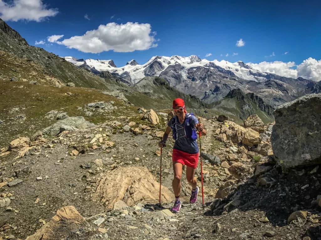 Female trail runner with red skirt and headband and two poles in rocky terrain with snowy mountains in the background