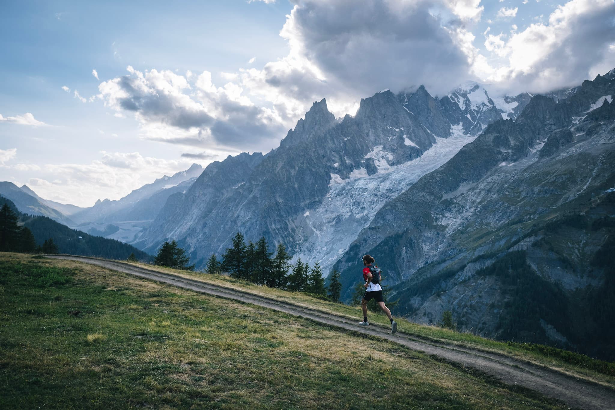 Runner runs up hill on grassy trail with alpine mountains in background