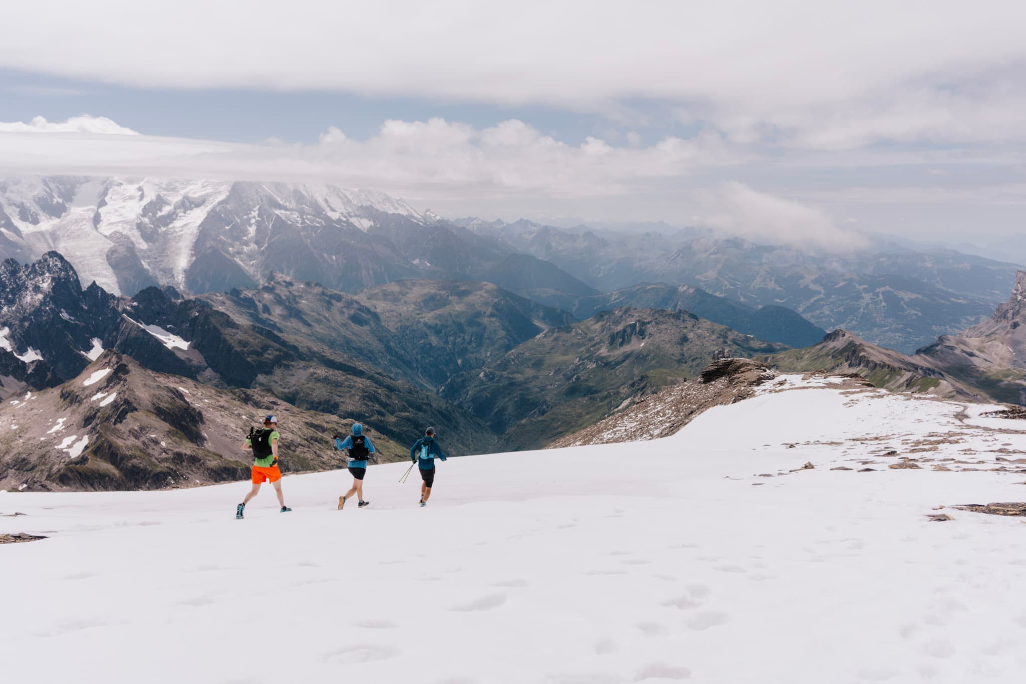 Three runners run downhill on the snow with Alpine mountains in background