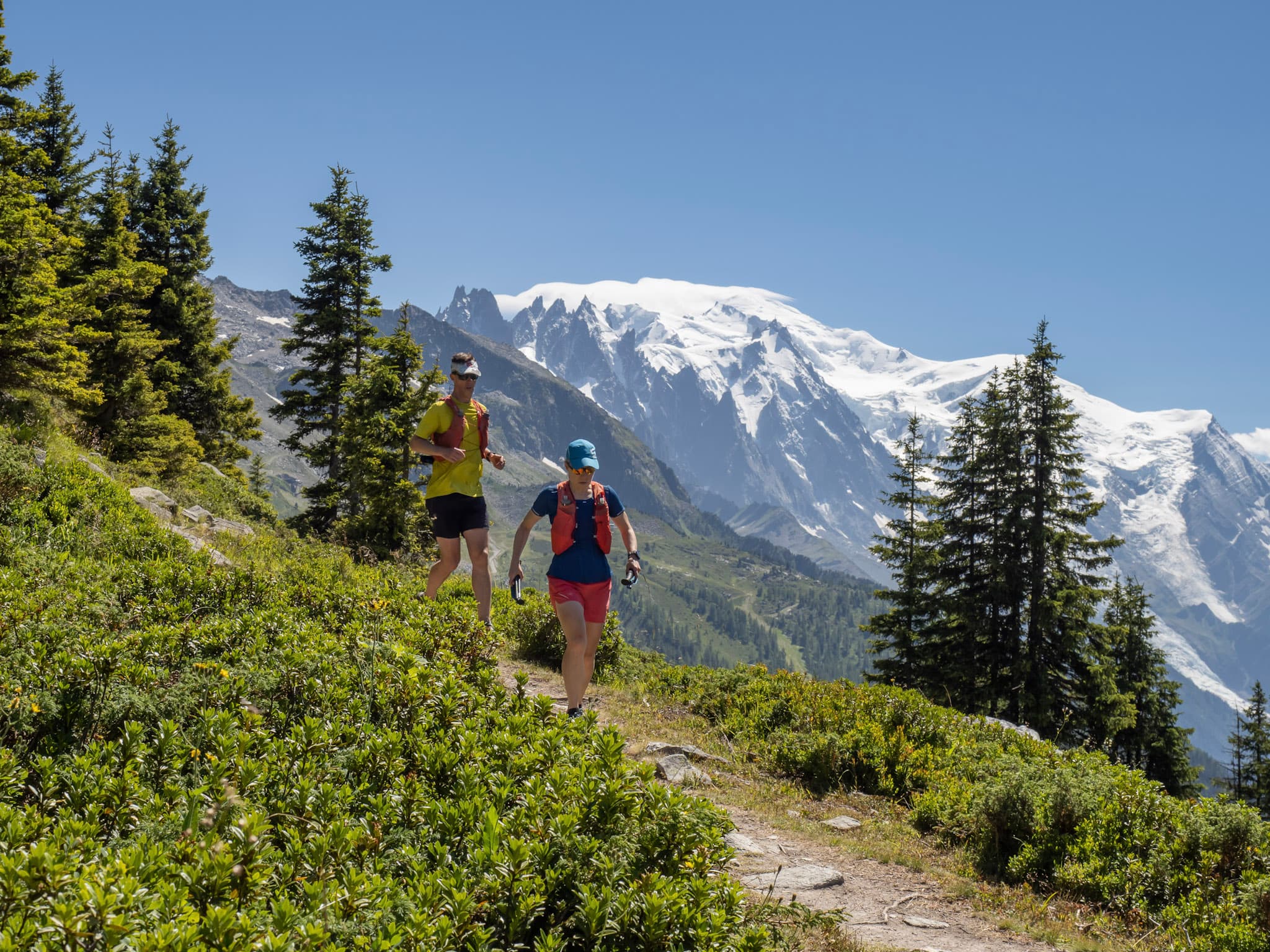 2 runners run along trail with alpine mountains in the background