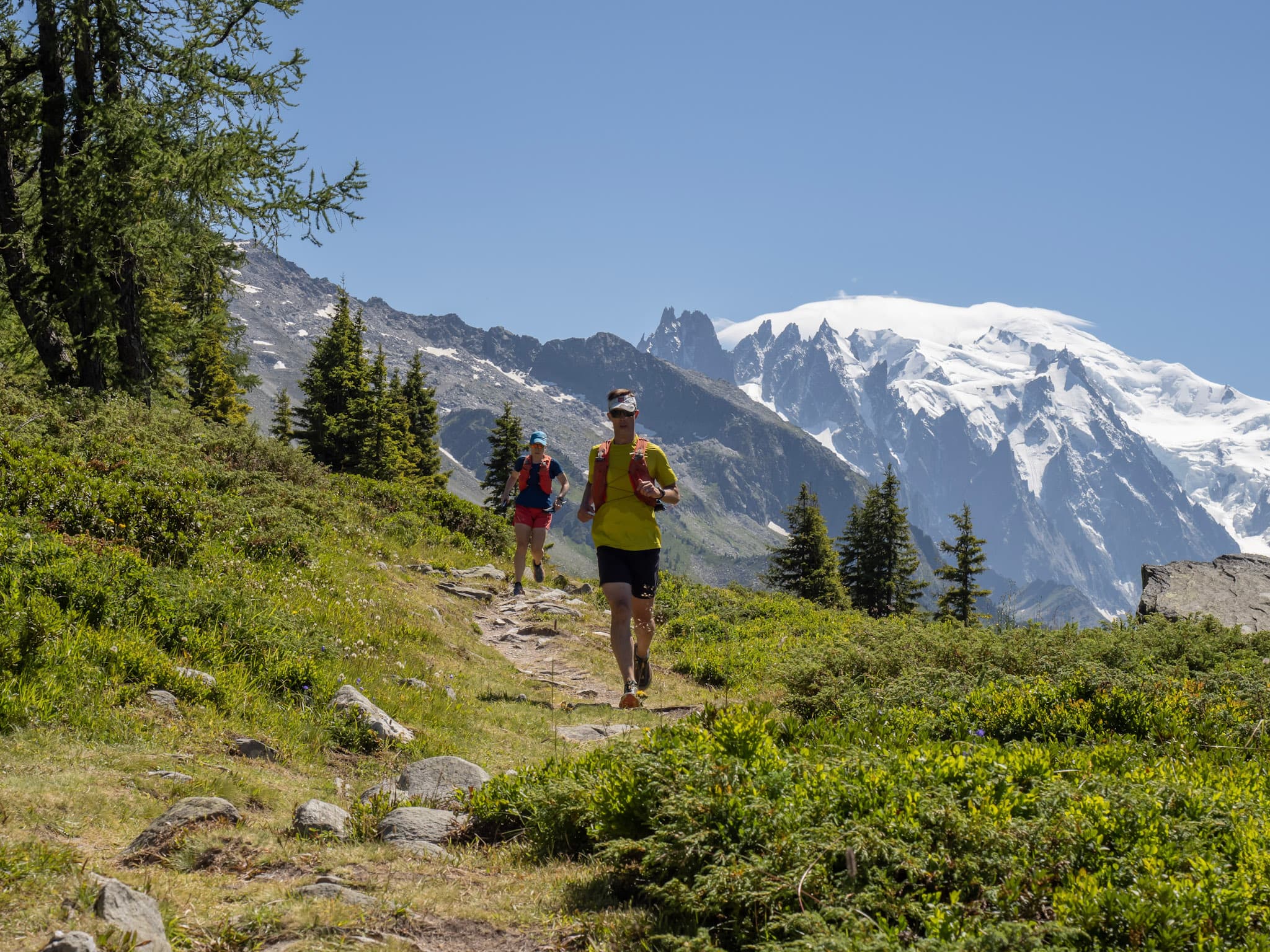 2 runners run along trail with alpine mountains in the background