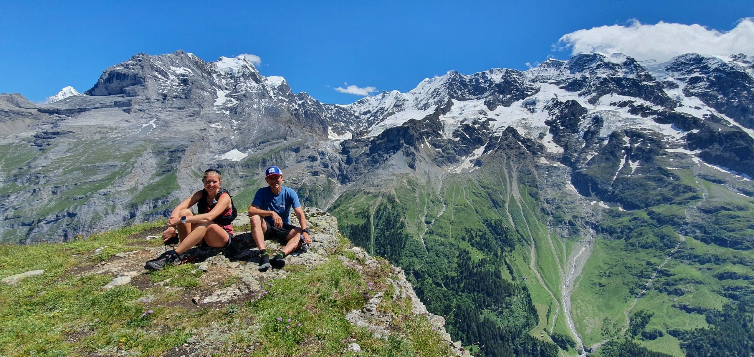 Male and female runner sitting on grass and rock high above Stechelberg with green, rocky and snowy mountains at the back