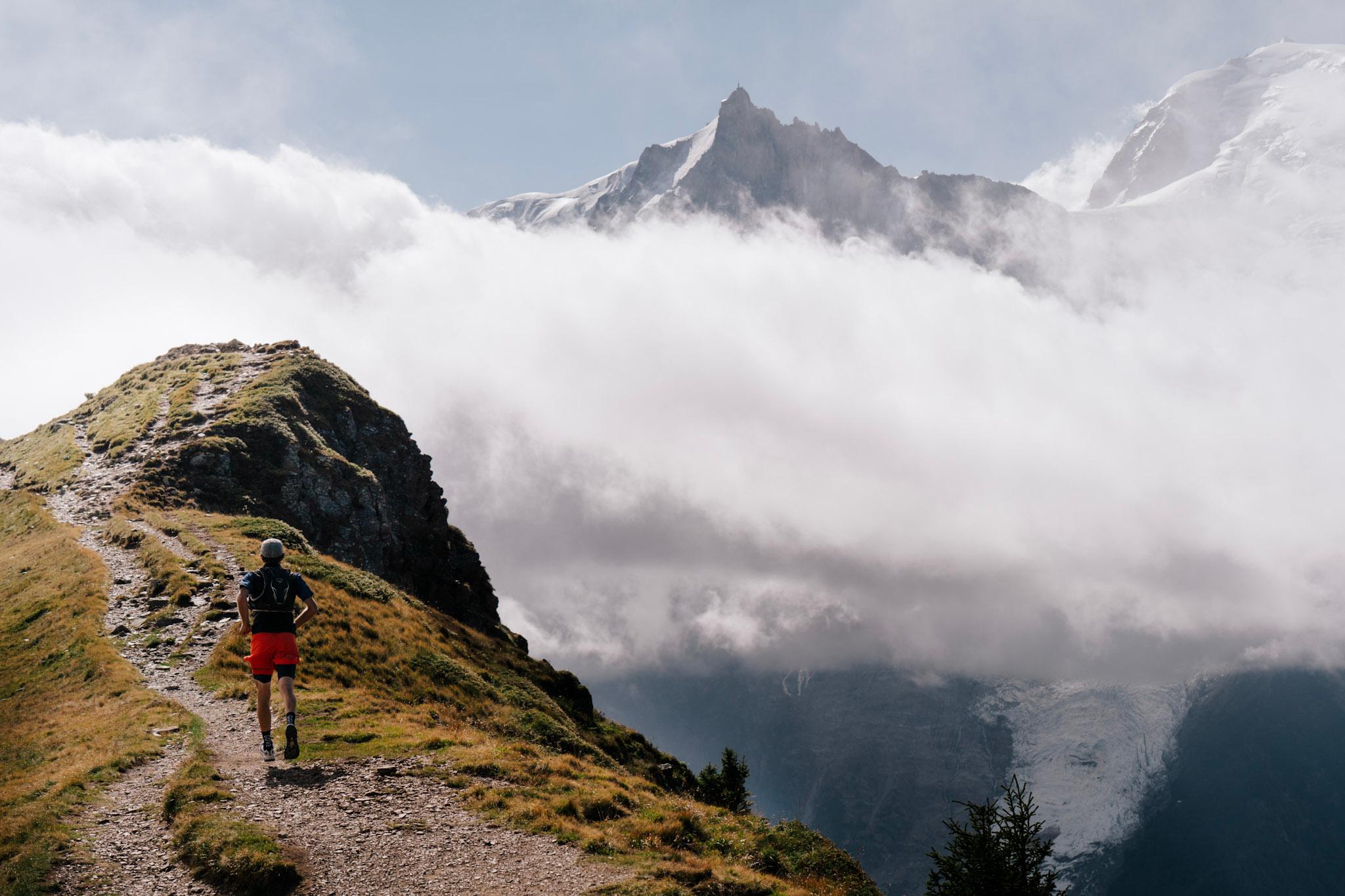 Runner runs along ridgeline Trail with Alpine Mountains and clouds in background