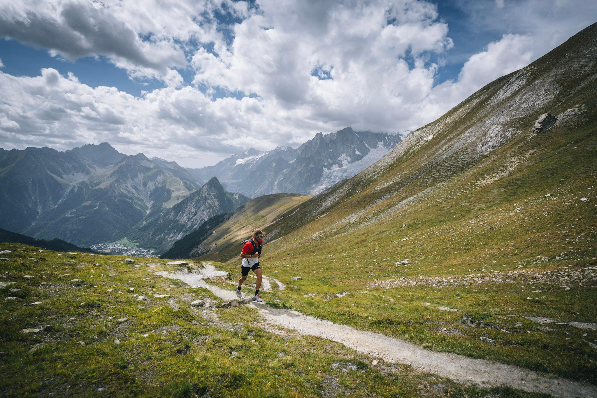 Runner runs up hill on grassy trail with alpine mountains in background