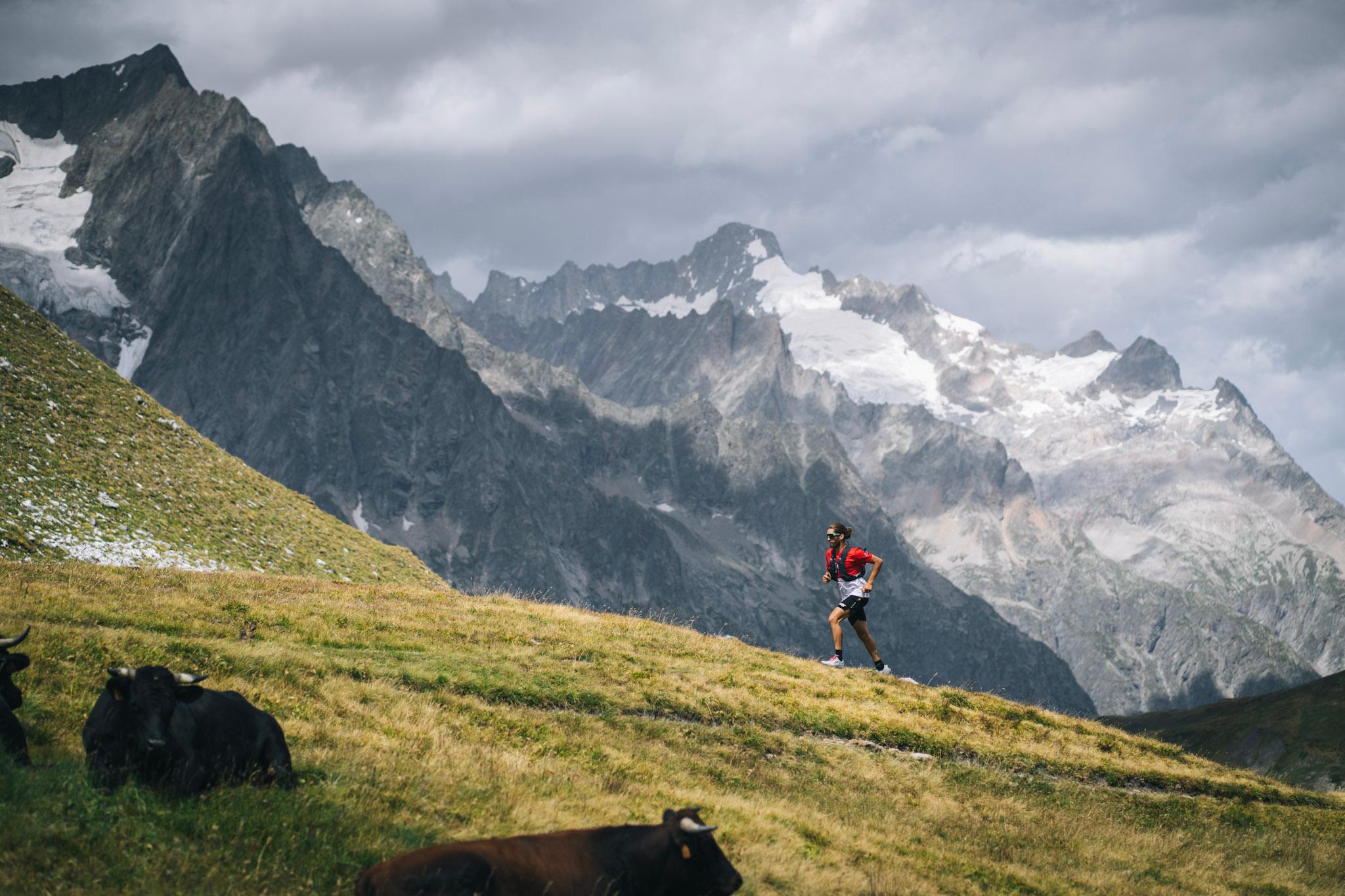 Runner runs up hill on grassy trail with alpine mountains in background