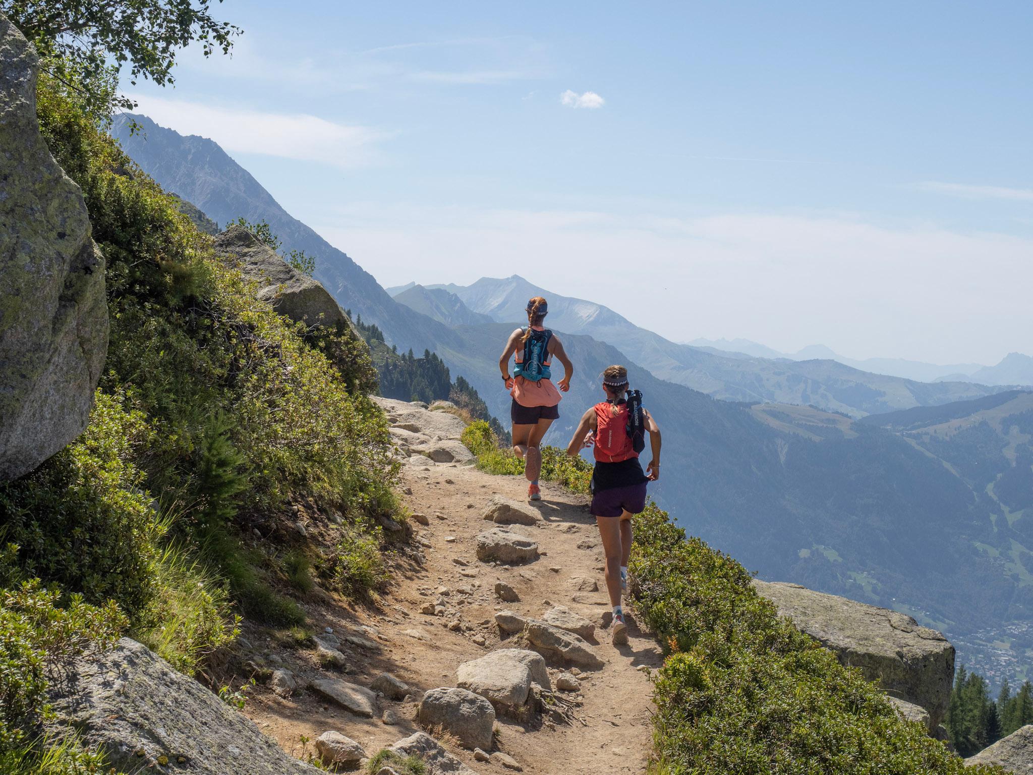 Two runners run around corner on high mountain traversing Trail with green mountains in the background