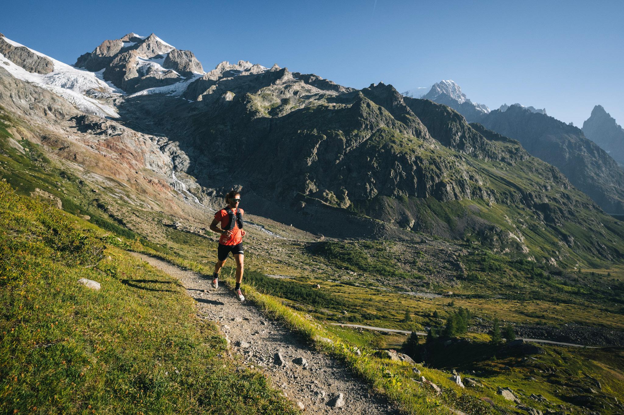 runner runs down trail with alpine mountains and meadows in background