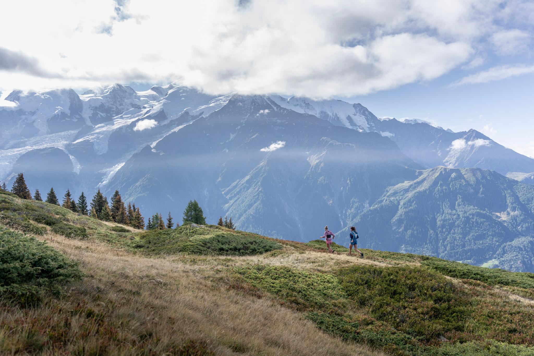 2 runners in the distance running through high alpine meadows with alpine mountains in background