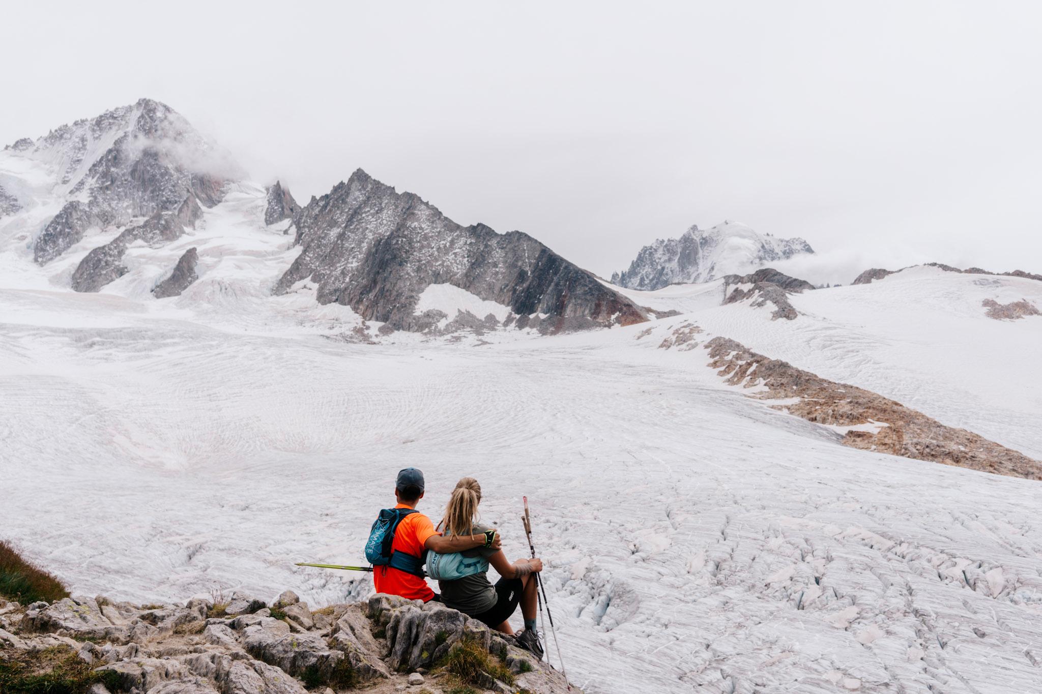 2 runners sit by glacier looking at alpine mountains