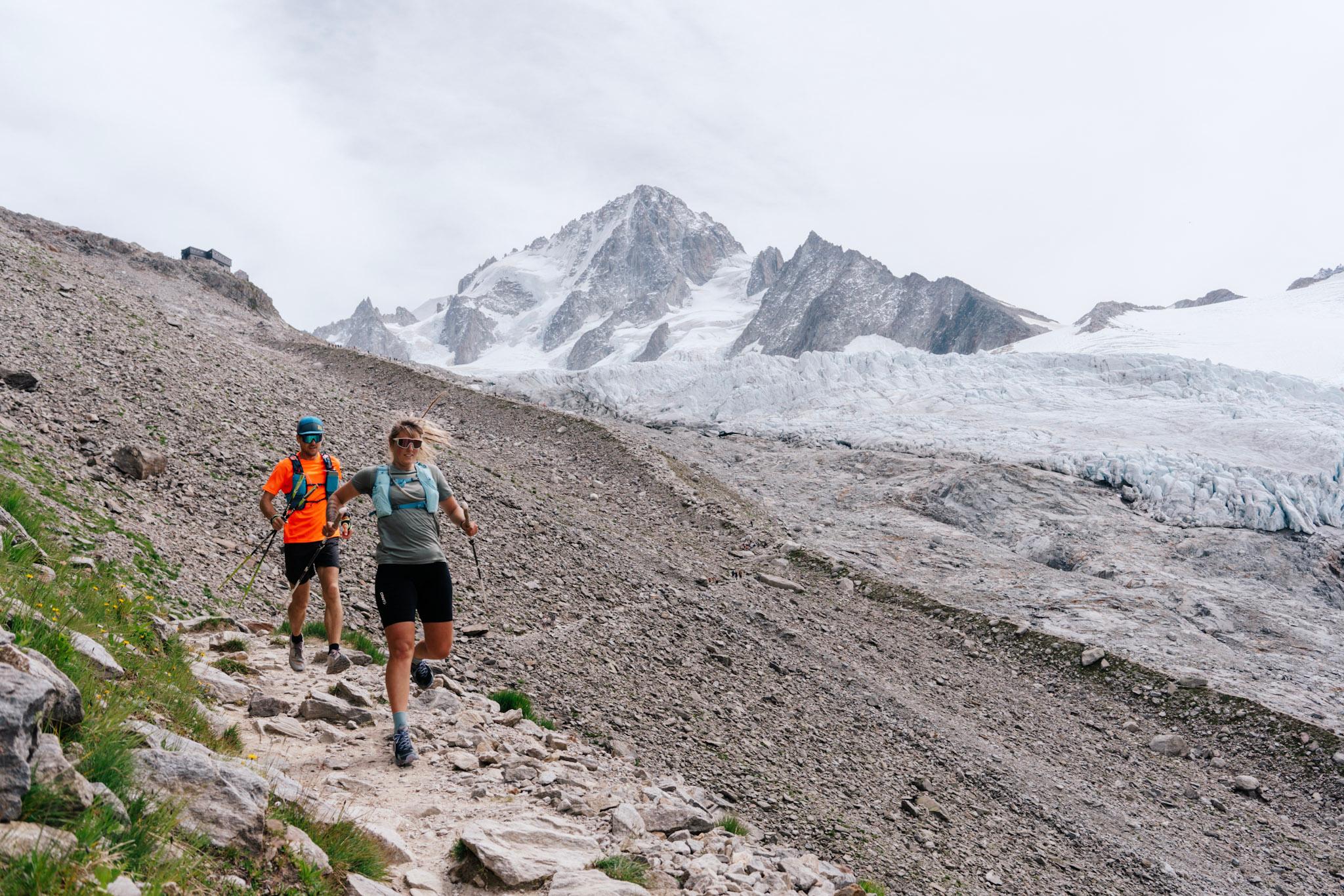 2 runners run down rocky trail with alpine mountains behind