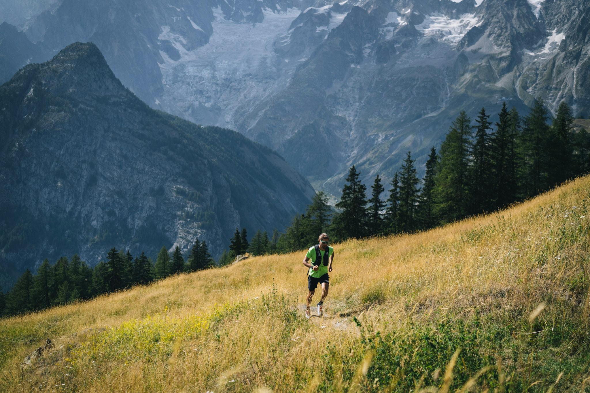 Runner runs single track through grassy frail with forest and mountains in the background