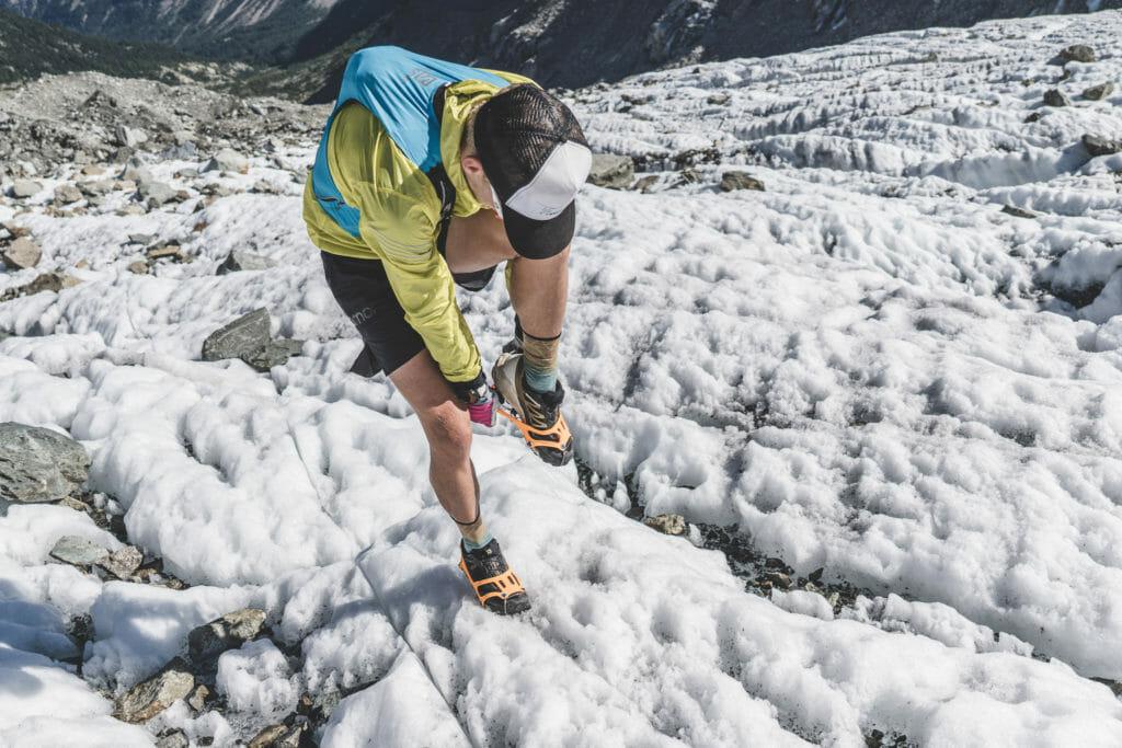 Trailrunner with yellow jacket and blue trail run vest putting micro-spikes on to cross glacier