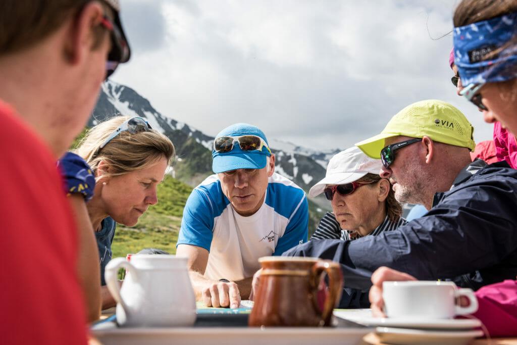 Guide showing trails on a map to trail runners who drink coffee at a table outside a mountain hut in Courmayeur