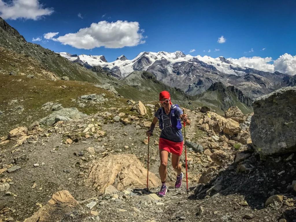Female trail runner with red skirt and headband and two poles in rocky terrain with snowy mountains in the background