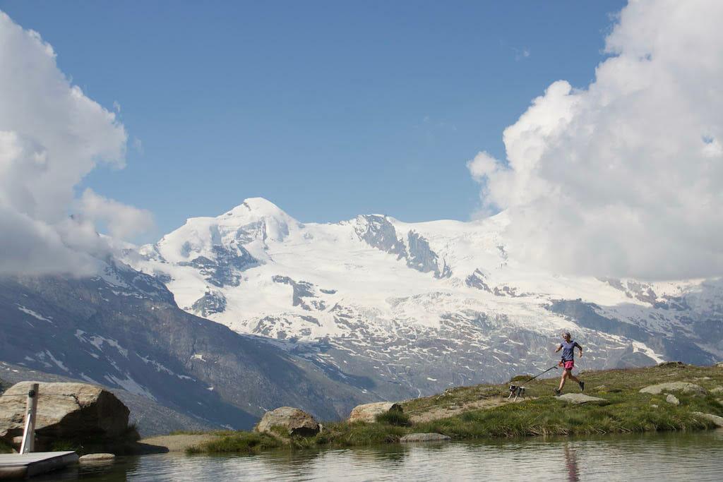 Siebrig Scheeres and her dog running along a mountain lake with snowy mountains at the back and some clouds