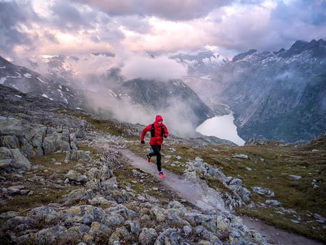 Urs Baumgartner with red jacket running on a single trail at the Grimselpass above a lake and clouds
