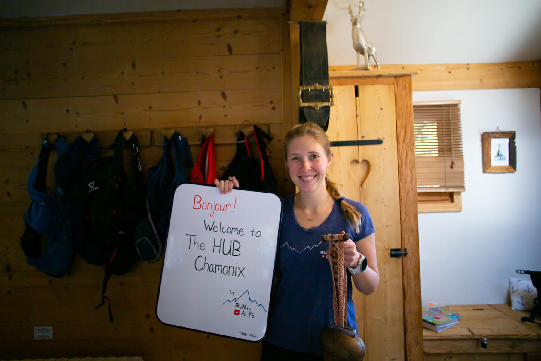 Blond female with handwritten "Bonjour! Welcome to the HUB Chamonix" sign and cow bell
