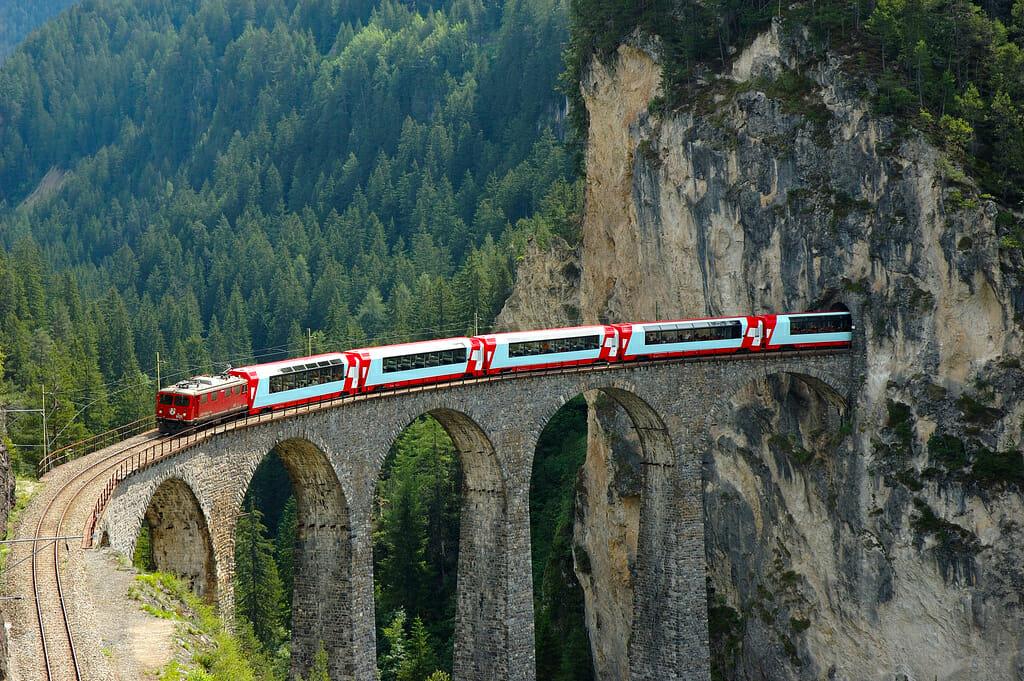 Red and white train with big windows on a high ancient pillar bridge going into a tunnel in the rocks