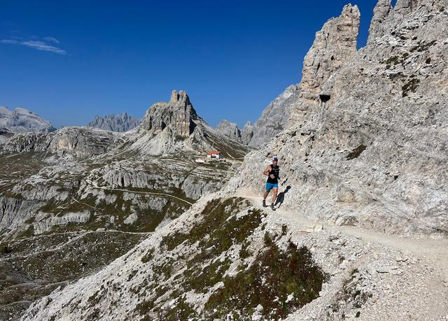 A man trail running in the Dolomites with a mountain hut in the background.